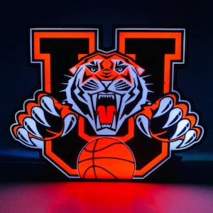 May include: A glowing orange and black sign featuring a tiger head with a basketball. The tiger is inside a letter "U" with sharp claws and an open mouth. The basketball is orange and the background is dark.