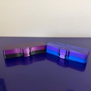 May include: Two rectangular, hinged boxes. One is purple, black, and pink. The other is blue and purple with a geometric design on the lid. Both have a clasp closure. The boxes are on a reflective, dark blue surface.