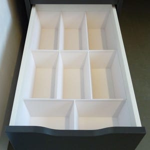 Puede incluir: Un organizador de cajones con múltiples compartimentos para organizar artículos pequeños. El organizador está hecho de plástico blanco y cabe dentro de un cajón.