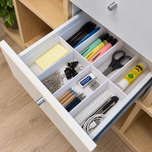 May include: A white desk drawer with a grid organiser containing office supplies including black and blue pens, pastel highlighters, black binder clips, silver paper clips, a white eraser, a glue stick, pencils, a ruler, a utility knife, and a cable.