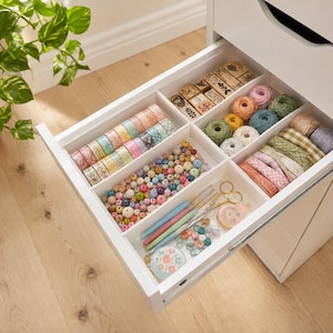 Puede incluir: Un mueble de almacenamiento blanco con un cajón abierto que contiene materiales de manualidades. El cajón tiene separadores con rollos de cinta decorativa, sellos de madera, ovillos de lana, cuentas, ganchillos, tijeras y botones variados.