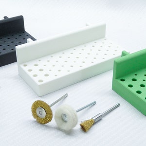 Op de afbeelding: Drie verschillende gekleurde plastic opbergorganisatoren met een raster van gaten om kleine gereedschappen in te bewaren. De organisatoren zijn zwart, wit en groen. Er zijn ook verschillende kleine metalen gereedschappen en een paar kleine borstels.