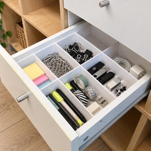 Op de afbeelding: Een witte bureaulade met een vakverdeling met kantoorbenodigdheden, waaronder zilveren paperclips, zwarte binderclips, gekleurde memoblaadjes, pennen, rollen washi-tape, USB-sticks, een witte oplaadkabel en een kleine witte stroomadapter.