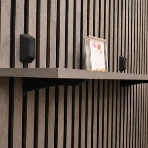 Op de afbeelding: Een modern wandrek met een grijze afwerking, ondersteund door zwarte beugels, is gemonteerd tegen een verticale houten lattenwand. Het rek bevat een ingelijste foto met hartvormige ontwerpen en twee kleine, zwarte, rechthoekige luidsprekers.