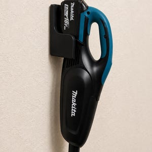 Könnte beinhalten: Schwarzer und türkisblauer kabelloser Handstaubsauger an der Wand. Der Staubsauger hat den Markennamen "Makita" auf dem Gehäuse aufgedruckt. Der Akku ist mit "5.0Ah 18V" beschriftet.