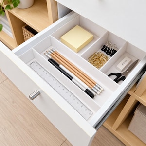 May include: A white desk drawer organiser with multiple compartments containing a clear ruler, four wooden pencils, a black pen, a stack of yellow sticky notes, gold paper clips, two black binder clips, a white eraser labelled MONO, and scissors.