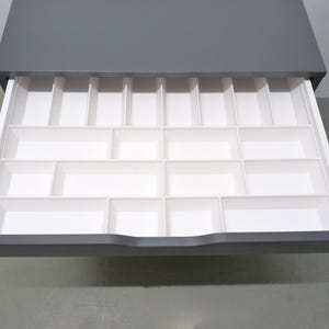 May include: A dark grey drawer pulled open to reveal a white plastic organiser insert. The insert features multiple rectangular and square compartments of varying sizes, designed for sorting small items within the drawer space.