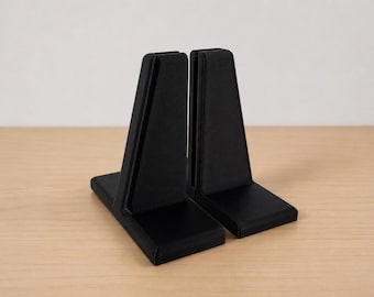 May include: A pair of black bookends featuring a solid L-shaped base and a vertical, slightly tapered upright support. The matte black finish covers the entire surface of both pieces, which are positioned side-by-side to show their structure.