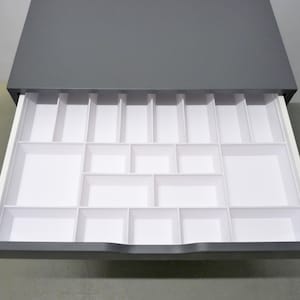 Könnte beinhalten: Eine dunkelgraue Schublade mit einem weißen Kunststoffeinsatz, der über mehrere rechteckige und quadratische Fächer zur organisierten Aufbewahrung verfügt. Die Schublade ist geöffnet und zeigt die gitterartige Anordnung des Einsatzes.