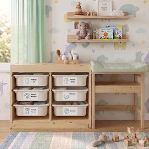 May include: A light wood nursery storage unit with six white plastic bins labeled with German text and icons for baby clothing and supplies, positioned next to a matching changing table with a light green pad and two wall-mounted shelves above.