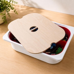 Puede incluir: Una bandeja de almacenamiento rectangular blanca con interior rojo, que cuenta con una tapa de madera clara con una abertura ovalada central. La bandeja está parcialmente cubierta por la tapa, dejando ver pequeños bloques de construcción.