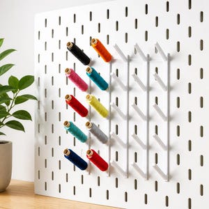 May include: A white pegboard with a grid of holes and white hooks, displaying colourful spools of thread. The thread colours include black, orange, pink, teal, red, yellow, grey, and navy blue. A potted plant is visible on the left.