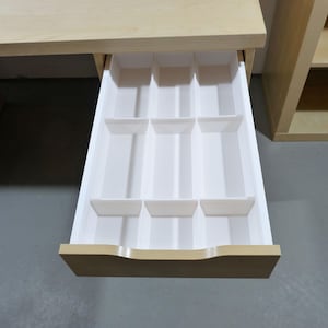 Könnte beinhalten: Ein helles Holzschreibtisch mit einer geöffneten Schublade, die einen weißen Kunststoff-Organizer zeigt. Der Organizer hat mehrere Fächer unterschiedlicher Größe. Der Schreibtisch ist Teil eines größeren Möbelsets, mit einem passenden Schrank im Hintergrund.