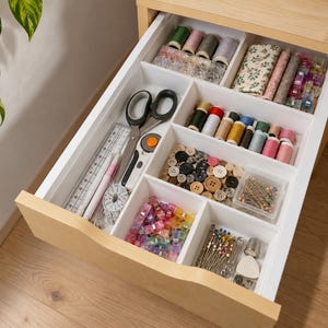 Op de afbeelding: Een lichte houten lade met witte verdelers met naaibenodigdheden zoals klossen garen, stofresten, een schaar, een rolsnijder, een doorzichtige liniaal, een meetlint, diverse knopen, naaiklemmen, spelden en een metalen vingerhoed.