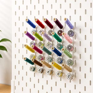 Könnte beinhalten: Ein weißes Pegboard-Organizer mit farbenfrohen Garnrollen und Spulen. Die Rollen sind in Blau-, Rot-, Lila-, Grün-, Rosa-, Gelb- und Brauntönen gehalten. Die Spulen sind silber- und goldfarben. Eine Aufbewahrungslösung für Näh- und Bastelbedarf.