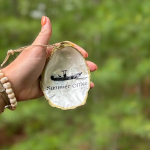 May include: An oyster shell ornament featuring a black boat illustration and the text "Summer Office." The shell has a pearlescent interior and a gold-toned edge. A twine loop is attached for hanging.