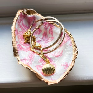 May include: A decorative oyster shell jewelry dish with a gold-painted rim and a pink floral pattern interior. The dish holds gold hoop earrings and a gold seashell pendant necklace, perfect for storing small accessories.