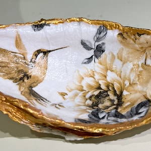 Peut inclure: Un plat en céramique blanche avec un bord doré, orné d'un colibri et d'un motif floral peint. Le plat est parfait pour ranger des bijoux, des clés ou d'autres petits objets.