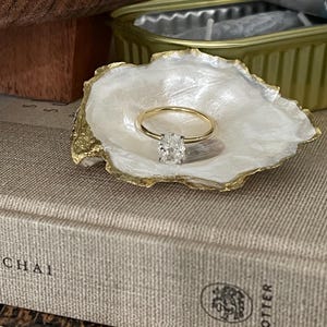 Puede incluir: Un anillo de oro con un gran diamante ovalado transparente colocado en un porta anillos con forma de concha blanca.