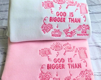 Puede incluir: Dos sudaderas dobladas, una blanca y otra rosa, con texto y gráficos bordados. El texto dice "GOD IS BIGGER THAN" con ilustraciones circundantes. La sudadera rosa está debajo de la blanca.