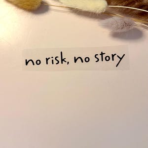Könnte beinhalten: Ein transparenter Aufkleber mit dem schwarzen Text no risk, no story in einer handschriftlichen Schriftart. Der Aufkleber ist unter einem Bündel getrockneter, flauschiger und federartiger botanischer Stängel in Beige, Hellblau und Creme platziert.