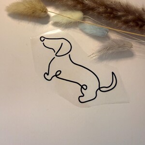 Könnte beinhalten: Eine schwarze Strichzeichnung eines Dackelhundes auf einem transparenten Aufkleber. Der Hund ist in einem minimalistischen Stil dargestellt, wobei eine einzige durchgehende Linie seine Kontur bildet. Der Aufkleber befindet sich auf einer weißen Oberfläche, mit getrockneten Ziergräsern im Hintergrund.