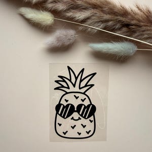 Pode incluir: Um autocolante de vinil preto com um ananás de desenho animado com um topo de folhas, uma pequena boca sorridente e óculos de sol em forma de coração. O design está impresso numa folha de suporte retangular transparente.