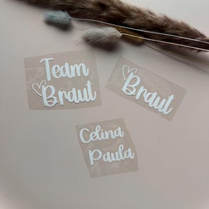 Może przedstawiać: Trzy białe naklejki winylowe na r&oacute;żowej powierzchni. Naklejki głoszą "Team Braut", "Braut" i "Celina Paula" w czcionce skryptowej. Suszona trawa ozdobna w tle.