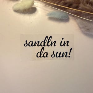 Peut inclure: Un autocollant transparent rectangulaire présentant le texte noir en cursive sandln in da sun! avec un point d'exclamation. Le lettrage est disposé sur deux lignes, la première ligne indiquant sandln in et la seconde ligne da sun!.