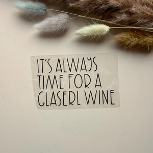 Peut inclure: Un autocollant transparent avec le texte "IT'S ALWAYS TIME FOR A GLASERL WINE" en noir, dans une police stylisée. L'autocollant est placé sur un fond clair, avec des plantes décoratives séchées en haut.