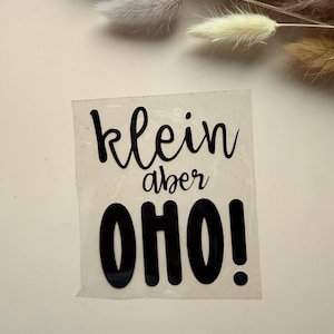 Könnte beinhalten: Ein transparenter Aufkleber mit dem schwarzen Text "klein aber OHO!" in einer Kombination aus Schreibschrift und fetter serifenloser Schrift. Die Wörter "klein" und "aber" sind in einer dünnen Schriftart gehalten, während "OHO!" in großen, fetten Buchstaben steht.