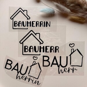 Può includere: Decalcomanie in vinile adesivo trasparente con testo nero e illustrazioni di case. Le decalcomanie presentano le parole "BAUHERRIN" e "BAUHERR". Alcune includono un design a cuore e camino. Su una superficie rosa chiaro.