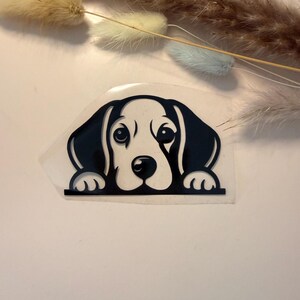 Könnte beinhalten: Ein schwarz-weißer Aufkleber mit einem Cartoon-Hund, der über eine horizontale Linie schaut. Der Hund hat große, ausdrucksstarke Augen und Schlappohren. Der Aufkleber befindet sich auf einer transparenten Rückseite.