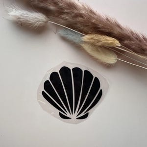 Könnte beinhalten: Ein schwarzer Muschelschalen-Aufkleber auf weißem Hintergrund. Der Aufkleber hat die Form einer Jakobsmuschel. Getrocknetes Ziergras in Braun-, Beige- und Blautönen befindet sich im Hintergrund.
