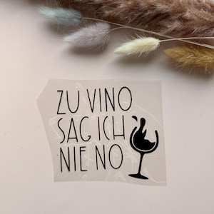 Può includere: Un adesivo trasparente con il testo nero ZU VINO SAG ICH NIE NO e un grafico di un bicchiere di vino con uno schizzo. L'adesivo è presentato su una superficie chiara con piume di erba secca posizionate sul bordo superiore dell'immagine.