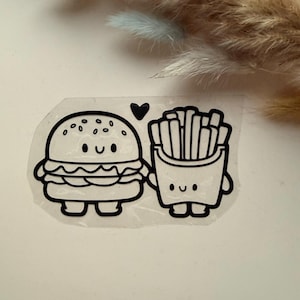 May include: A black and white sticker depicting a cartoon burger and a carton of chips, both with smiling faces. A small black heart is above the burger. The sticker is on a clear background.