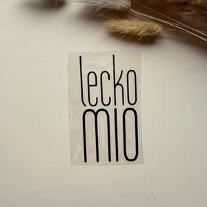 Może przedstawiać: Przezroczysta naklejka z napisem "lecko mio" czarną, minimalistyczną czcionką. Naklejka jest prostokątna i znajduje się na jasnej powierzchni, z suszonymi akcentami kwiatowymi w tle.
