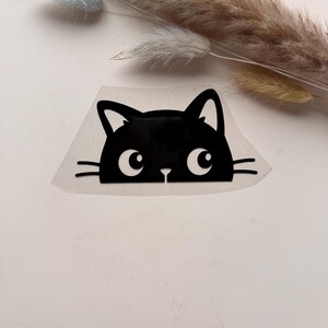 Könnte beinhalten: Ein schwarzer Vinyl-Aufkleber, der den Kopf einer Katze mit großen weißen Augen und dünnen Schnurrhaaren zeigt. Das Katzendesign ist in einem flachen, grafischen Stil mit spitzen Ohren und einer kleinen Nase auf einer transparenten Trägerfolie.