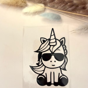Könnte beinhalten: Schwarzer und weißer Cartoon-Einhorn-Aufkleber mit Sonnenbrille. Das Einhorn hat ein Horn, eine fließende Mähne und sitzt. Das Bild befindet sich auf einem transparenten, rechteckigen Aufkleber.