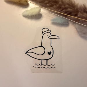 Könnte beinhalten: Ein schwarz-weißer Aufkleber mit einer Cartoon-Möwe, die einen Hut und ein Herz trägt. Die Möwe steht im Wasser, dargestellt durch einfache Wellenlinien. Der Aufkleber befindet sich auf weißem Hintergrund.