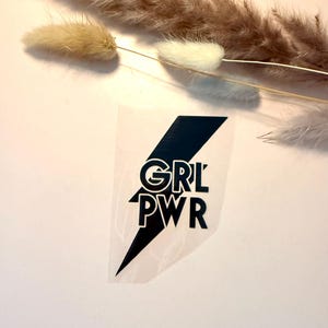Könnte beinhalten: Ein schwarzer Aufkleber in Blitzform mit dem weißen Text GRL PWR, der mittig in der Form platziert ist. Das Design ist auf einer transparenten Klebefolie gedruckt und zeigt kräftige, klare Linien sowie eine scharfe, kantige Silhouette.