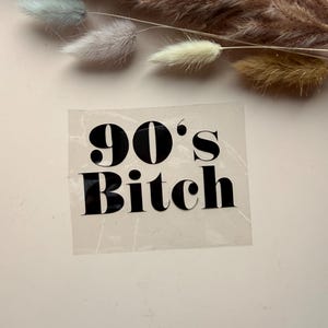 Peut inclure: Un autocollant transparent avec le texte "90's Bitch" en gras, en noir. L'autocollant est placé sur une surface rose clair, avec de l'herbe ornementale séchée en arrière-plan.