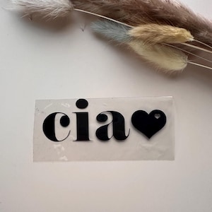 Peut inclure: Un autocollant transparent avec le mot "ciao" en noir gras, accompagné d'un symbole de cœur noir. L'autocollant est rectangulaire et posé sur une surface blanche. De l'herbe ornementale séchée est en arrière-plan.