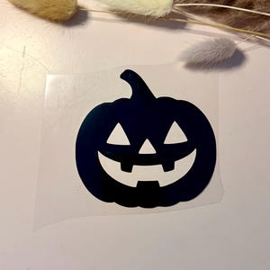 Könnte beinhalten: Ein schwarzer Vinyl-Aufkleber mit einem Kürbis-Design, das einen Stiel, zwei dreieckige Augen, eine kleine dreieckige Nase und ein breites, zahniges Grinsen aufweist. Der Aufkleber befindet sich auf einer transparenten, rechteckigen Trägerfolie.