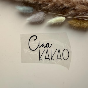 May include: A transparent sticker with the words "Ciao KAKAO" in black script and sans-serif font. The sticker is placed on a white surface, with dried flowers in the background.