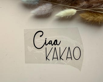 Może przedstawiać: Przezroczysta naklejka z czarnym napisem Ciao KAKAO wykonanym połączeniem czcionki pisanej i bezszeryfowej. Naklejka znajduje się pod wiązką suszonych, puszystych, jasnobrązowych i jasnoniebieskich łodyg ozdobnych traw.