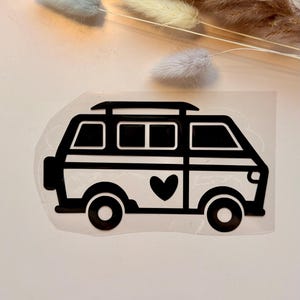 Könnte beinhalten: Ein schwarzer Vinyl-Aufkleber in Form eines stilisierten Campingbusses mit Dachgepäckträger, einem Reserverad am Heck und einem Herzsymbol auf der Seitenwand. Das Design ist auf einer transparenten Klebefolie mit klaren Linien dargestellt.