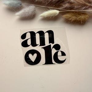 Könnte beinhalten: Ein schwarzer Vinyl-Aufkleber mit dem Wort "amore" in einer kräftigen, stilisierten Schriftart. Der Buchstabe "o" hat eine Herzform. Der Aufkleber befindet sich auf einer hellrosa Oberfläche, mit getrockneten Blumenstielen im Hintergrund.