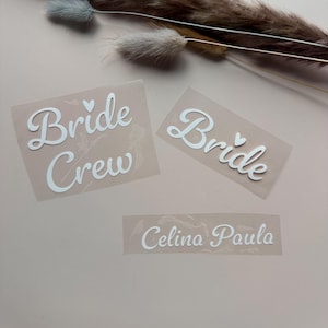 Könnte beinhalten: Drei weiße Wärmeübertragungs-Vinylaufkleber mit Text. Die Aufkleber lauten "Bride Crew", "Bride" und "Celina Paula". Die Aufkleber befinden sich auf einer hellrosa Oberfläche, mit getrockneten floralen Akzenten im Hintergrund.