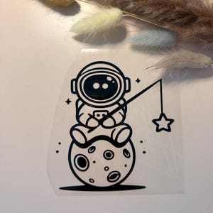 Può includere: Grafica in bianco e nero di un astronauta seduto su una luna, che pesca una stella. L'astronauta indossa un casco e tiene una canna da pesca con una stella alla fine. La luna ha crateri.